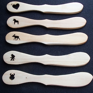 May include: A collection of five wooden spreaders with various etched designs. One features a heart cutout, while others display a horse, moose, a figure, and a bull. The spreaders are light-colored wood with a smooth finish.