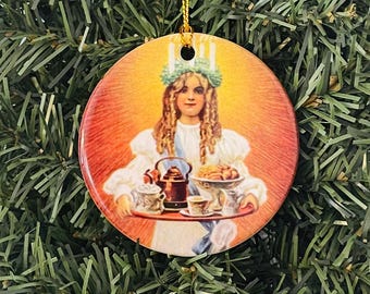 May include: Round Christmas ornament featuring a vintage illustration of a woman holding a tray with a teapot, cups, and pastries. The woman wears a crown of candles and a white dress. The ornament has a gold hanging string.
