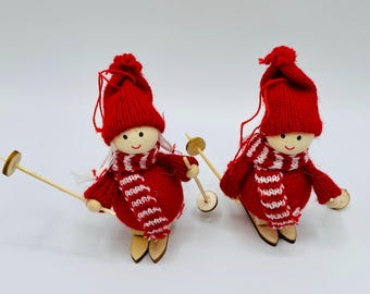 May include: Two handmade Christmas ornaments featuring wooden dolls dressed in red knitted outfits, hats, and striped scarves. Each doll holds wooden ski poles and skis. The ornaments are designed for holiday decor.