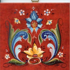 May include: Square, red decorative tile with a floral design. The design features a central blue flower with a yellow center, surrounded by blue and green leaves and smaller white and yellow flowers. The tile is framed with a gold metal hanger.
