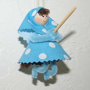 May include: A whimsical handmade doll with a wooden head, brown hair, and a smiling face. The doll wears a blue dress with white polka dots and a matching bonnet. It holds a wooden stick and has blue pipe cleaner arms and legs.