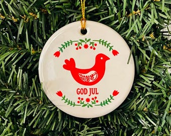 May include: Round white Christmas ornament with a red bird design and the words "GOD JUL". The ornament is decorated with red flowers and green leaves. A gold string is attached for hanging on a Christmas tree.