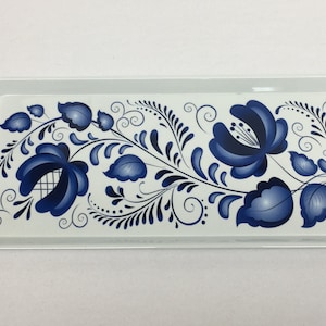 May include: A rectangular serving tray with a white background and a blue floral design. The tray features a repeating pattern of stylized blue flowers and leaves, with curved handles on each end.