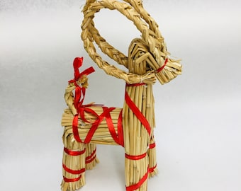 May include: A straw goat figurine with a braided head and large, curved horns. The goat is tied with red ribbon around its legs and body, and a red bow adorns its neck. This festive decoration is perfect for holiday displays.