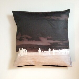 May include: A dark gray throw pillow with a white silhouette of a group of people walking across a desert landscape. The background is a dark gray with a light brown sky.