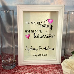 Könnte beinhalten: Weißer Rahmen mit einem Zitat darin: "Du bist mein Heute und all meine Morgen." Unter dem Zitat steht der Text "Sydney & Adam" und das Datum "28. Mai 2023".