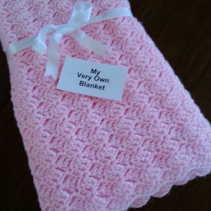 May include: A pink crocheted baby blanket with a white ribbon tied around it. The blanket has a label that says "My Very Own Blanket".