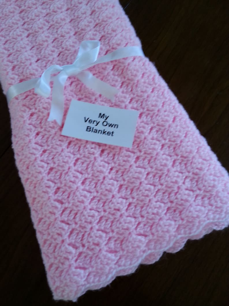 May include: A pink crocheted baby blanket with a white ribbon tied around it. The blanket has a label that says "My Very Own Blanket".