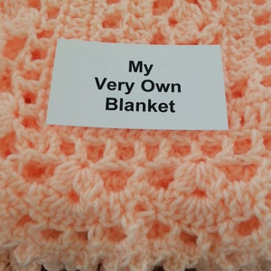 Puede incluir: Una manta de ganchillo de color melocot&oacute;n con una etiqueta blanca que dice "My Very Own Blanket".