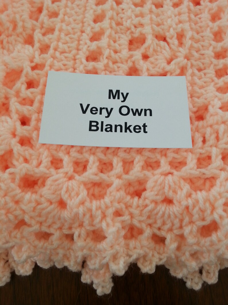 Puede incluir: Una manta de ganchillo de color melocot&oacute;n con una etiqueta blanca que dice "My Very Own Blanket".