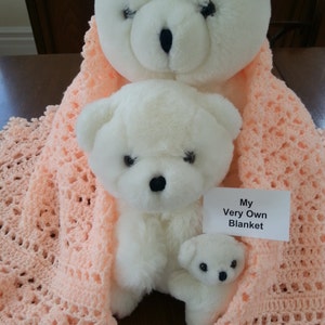 Puede incluir: Dos osos de peluche blancos, uno m&aacute;s grande que el otro, est&aacute;n acurrucados en una manta de ganchillo de color melocot&oacute;n. La manta tiene un delicado patr&oacute;n de encaje y el oso m&aacute;s peque&ntilde;o sostiene un peque&ntilde;o oso de peluche blanco. Una tarjeta blanca con el texto "My Very Own Blanket" es visible en la imagen.