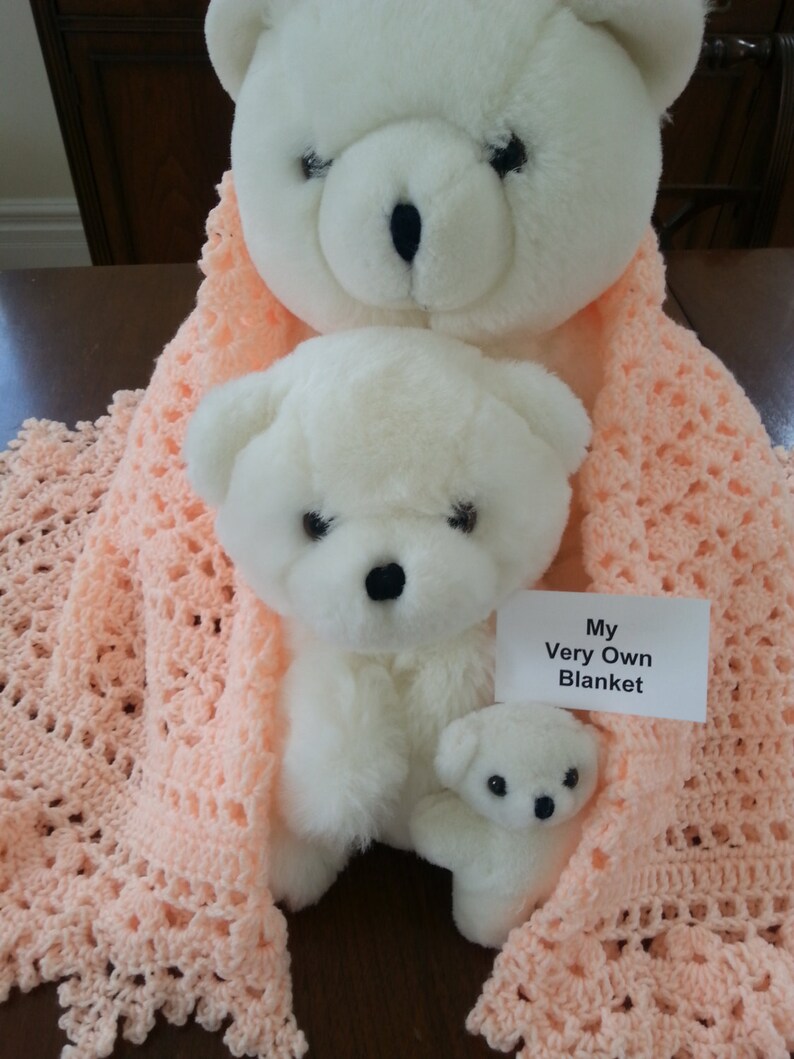 Puede incluir: Dos osos de peluche blancos, uno m&aacute;s grande que el otro, est&aacute;n acurrucados en una manta de ganchillo de color melocot&oacute;n. La manta tiene un delicado patr&oacute;n de encaje y el oso m&aacute;s peque&ntilde;o sostiene un peque&ntilde;o oso de peluche blanco. Una tarjeta blanca con el texto "My Very Own Blanket" es visible en la imagen.