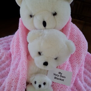 May include: Three white teddy bears of different sizes are nestled in a pink crocheted blanket. The largest bear is at the top, with the middle-sized bear in front of it. The smallest bear is in front of the middle-sized bear. A white tag with black text reads "My Very Own Blanket".
