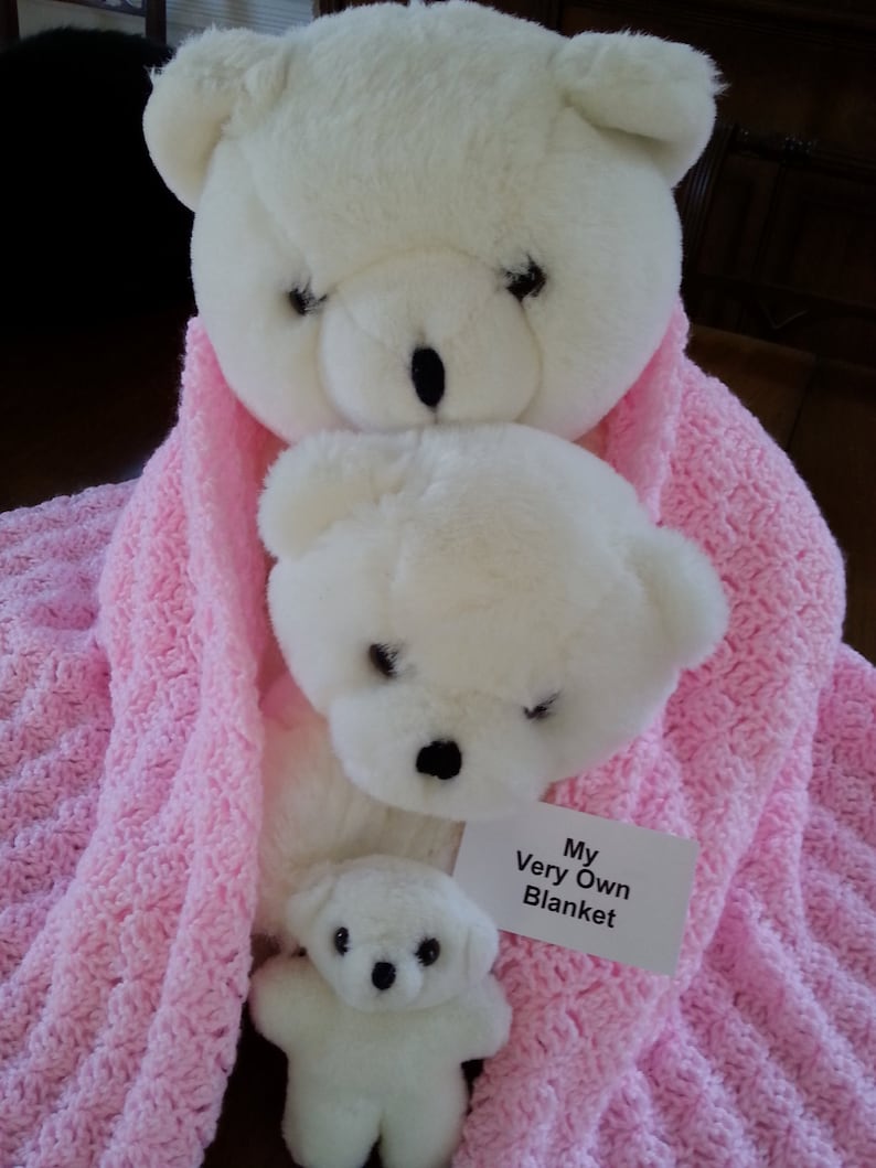 May include: Three white teddy bears of different sizes are nestled in a pink crocheted blanket. The largest bear is at the top, with the middle-sized bear in front of it. The smallest bear is in front of the middle-sized bear. A white tag with black text reads "My Very Own Blanket".