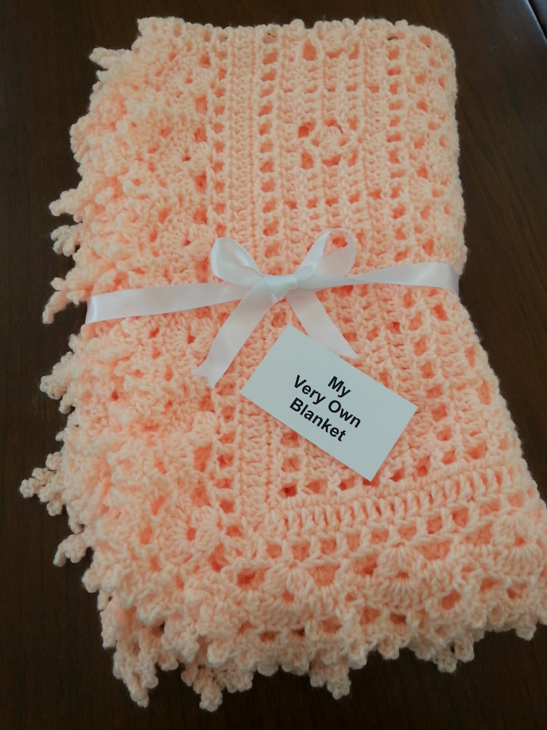 Puede incluir: Una manta de beb&eacute; de ganchillo de color melocot&oacute;n con una cinta de sat&eacute;n blanca y una etiqueta que dice "My Very Own Blanket".