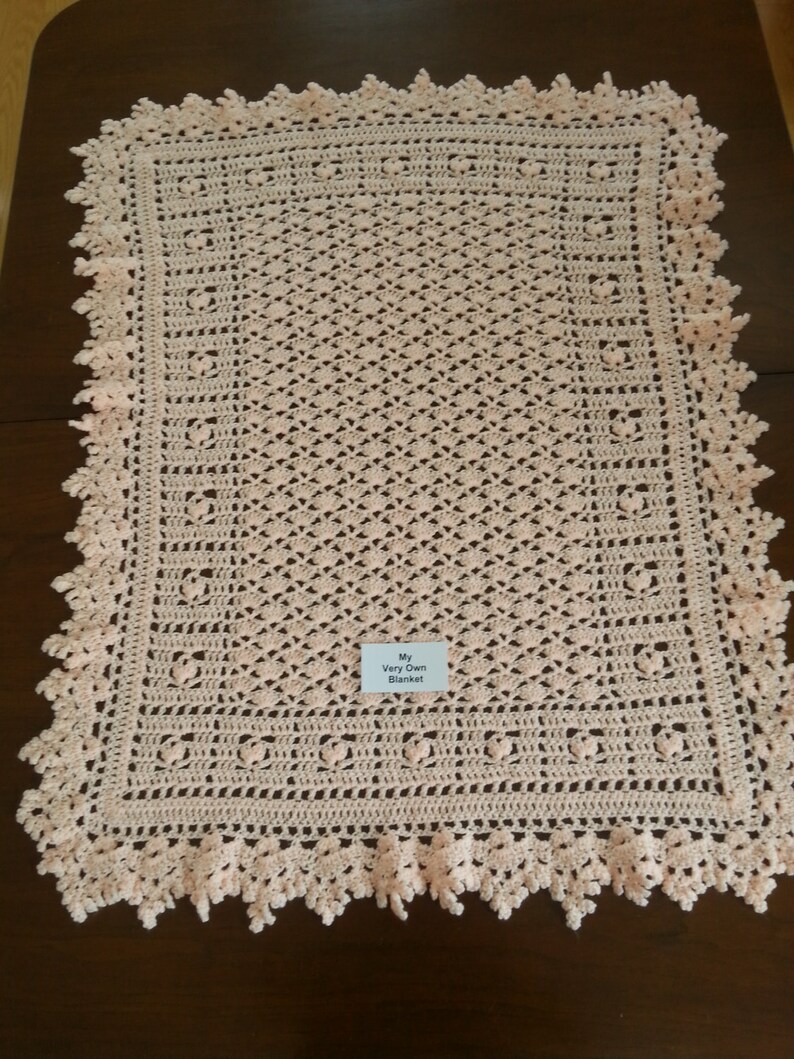 Puede incluir: Una manta de beb&eacute; de ganchillo en color rosa p&aacute;lido con un delicado borde de encaje. La manta tiene una etiqueta que dice "My Very Own Blanket".