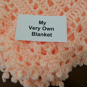 Puede incluir: Una manta de ganchillo de color melocot&oacute;n con una etiqueta que dice "My Very Own Blanket".