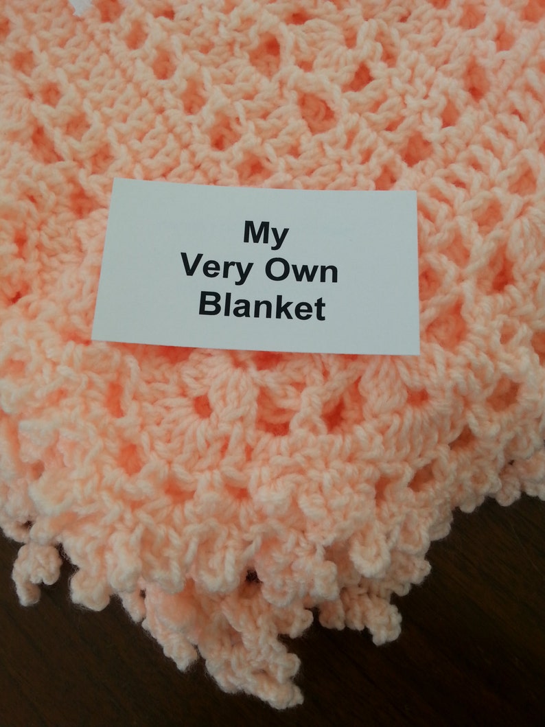 Puede incluir: Una manta de ganchillo de color melocot&oacute;n con una etiqueta que dice "My Very Own Blanket".