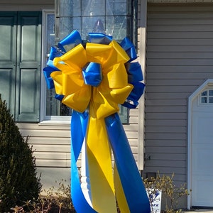 May include: A large yellow and blue bow tied around a white post. The bow is made of satin ribbon and is tied in a large, elaborate knot. The post is made of wood and is painted white.