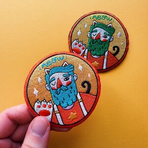 Puede incluir: Dos parches bordados circulares con un diseño de gato. Cada parche presenta un personaje de dibujos animados con barba, orejas de gato y una huella de pata. Los parches tienen un fondo dorado y la palabra "meow" encima de la cabeza del personaje.