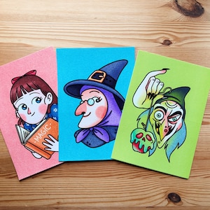 Puede incluir: Tres coloridas impresiones artísticas con ilustraciones caprichosas. Una muestra a una niña leyendo un libro con "MAGIC" en la portada, otra a una bruja con un sombrero y la última a un personaje de piel verde sosteniendo una manzana.