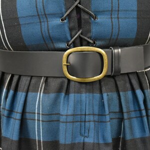 May include: A black leather belt with a large oval brass buckle. The belt is worn over a blue and black plaid skirt.