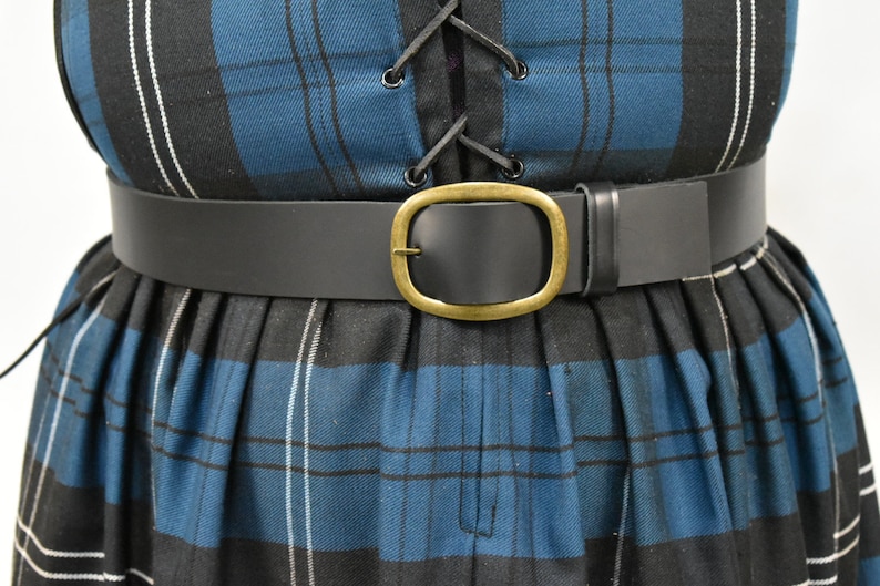 May include: A black leather belt with a large oval brass buckle. The belt is worn over a blue and black plaid skirt.