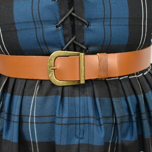 May include: A brown leather belt with a gold-toned buckle. The belt is worn over a blue and black plaid dress.