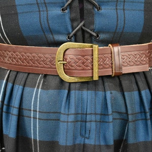 May include: A brown leather belt with a Celtic knot design and a brass buckle. The belt is worn over a blue and black plaid skirt.