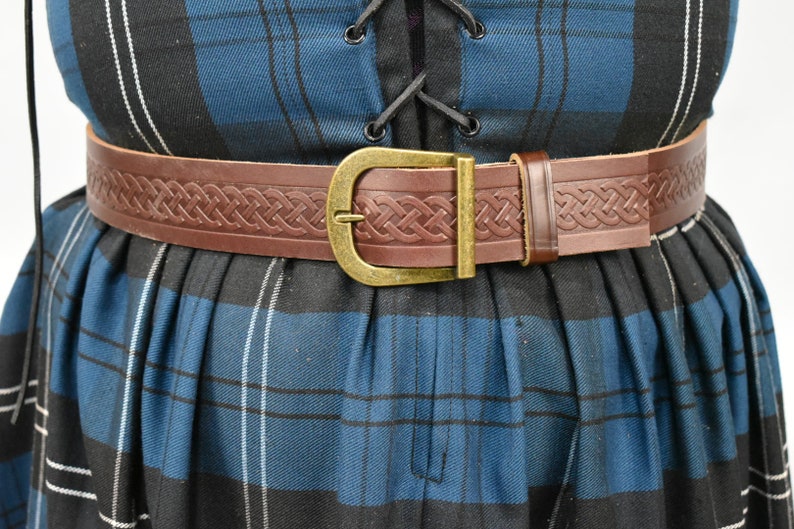 May include: A brown leather belt with a Celtic knot design and a brass buckle. The belt is worn over a blue and black plaid skirt.