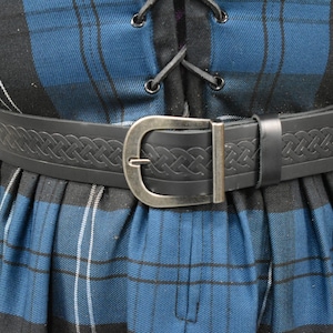 May include: A black leather belt with a Celtic knot design and a silver buckle. The belt is worn over a blue and black plaid skirt.