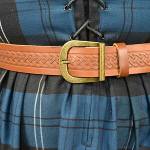 May include: A brown leather belt with a Celtic knot design and a brass buckle. The belt is worn over a blue and black plaid skirt.