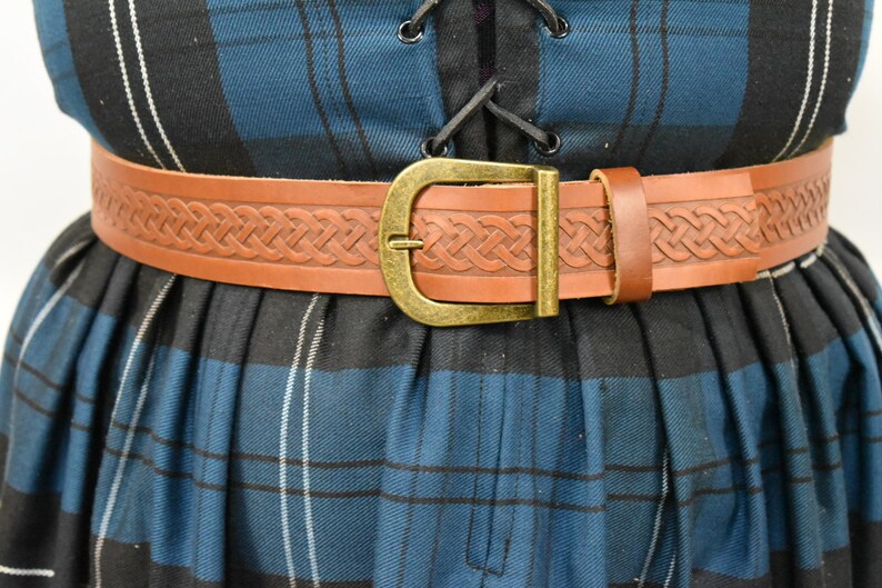 May include: A brown leather belt with a Celtic knot design and a brass buckle. The belt is worn over a blue and black plaid skirt.