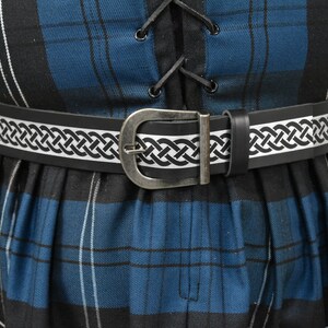 May include: A black leather belt with a silver buckle and a white Celtic knot design. The belt is worn over a blue and black plaid skirt.