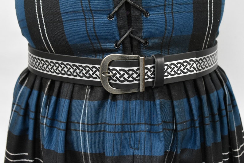 May include: A black leather belt with a silver buckle and a white Celtic knot design. The belt is worn over a blue and black plaid skirt.