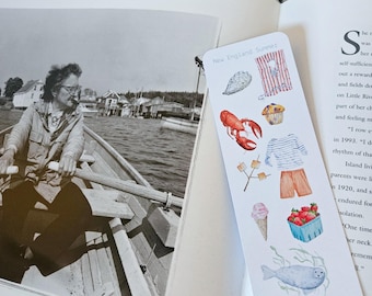 May include: A bookmark with the text "New England Summer" features watercolor illustrations of a lobster, ice cream cone, strawberries, and other summer-themed images. The bookmark is placed in an open book with a black and white photo.