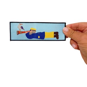 May include: Embroidered patch featuring a cartoon sailor lying down, with a seagull perched on his arm. The sailor wears a blue jacket, yellow pants, and a yellow hat. The patch has a light blue background and a black border.
