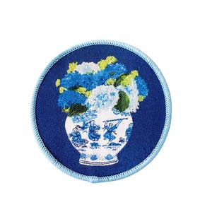 May include: A circular embroidered patch featuring a deep blue background with a light blue stitched border. The center displays a white vase decorated with blue figures, filled with a vibrant arrangement of blue, white, and green flowers.