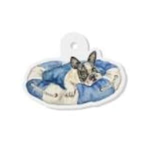 May include: A white, die-cut pet tag featuring a watercolor illustration of a French Bulldog resting in a blue and white dog bed. The tag has a hole at the top for attachment to a collar. The dog's fur is black and white.