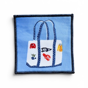 May include: Embroidered patch featuring a white tote bag with blue handles and accents. The bag is decorated with a red sweater, blueberries, a lobster, and a pennant that reads "MAINE". The patch has a blue border.