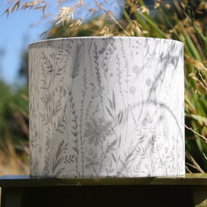 May include: A white lampshade with a grey floral pattern of various grasses and wildflowers. The lampshade is cylindrical and has a smooth, fabric texture.
