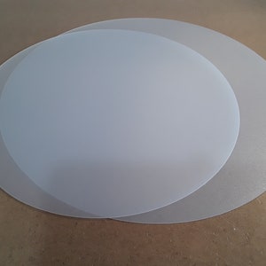 May include: Two white translucent round plastic sheets, one slightly larger than the other, on a brown surface.