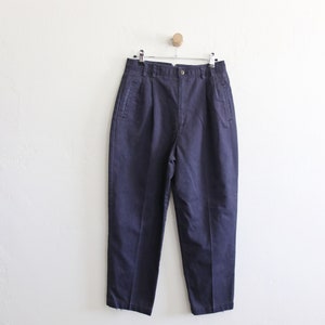 May include: A pair of dark blue pleated pants hanging on a hanger against a white wall.