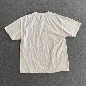May include: A cream-colored, short-sleeved t-shirt, laid flat on a gray surface. The shirt has a crew neck and appears to be made of a soft, lightweight fabric. The fabric has visible wrinkles, suggesting it has been worn or washed.