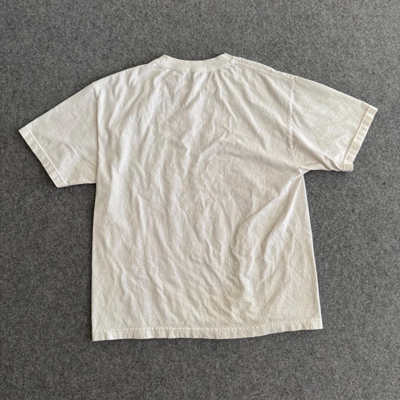 May include: A cream-colored, short-sleeved t-shirt, laid flat on a gray surface. The shirt has a crew neck and appears to be made of a soft, lightweight fabric. The fabric has visible wrinkles, suggesting it has been worn or washed.
