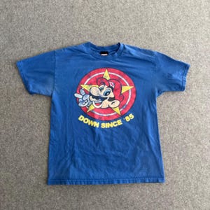 Könnte beinhalten: Blaues T-Shirt mit einem Vintage-Grafikdruck von Mario aus Super Mario Bros. Die Grafik zeigt Mario mit roter Mütze und Latzhose, einem gelben Stern und dem Text "Down Since '85".