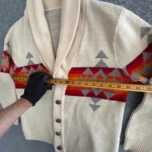 May include: A cream-colored knit cardigan with a shawl collar and a horizontal geometric pattern in red, orange, and grey. A wooden ruler is placed across the chest, showing a width measurement of approximately 22 inches from edge to edge.