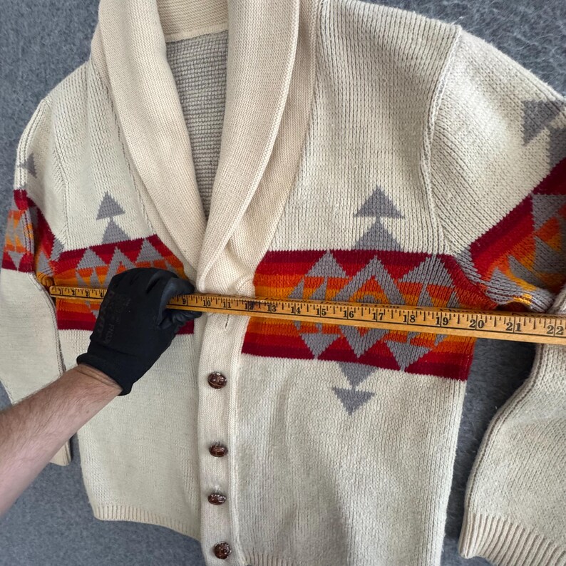 May include: A cream-colored knit cardigan with a shawl collar and a horizontal geometric pattern in red, orange, and grey. A wooden ruler is placed across the chest, showing a width measurement of approximately 22 inches from edge to edge.