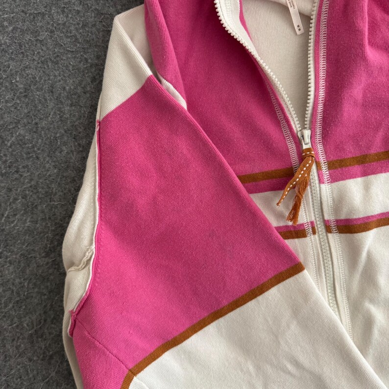 Op de afbeelding: Een hoodie met ritssluiting in een colorblock-ontwerp. De hoodie heeft een combinatie van witte, roze en bruine strepen. De rits heeft een bruine kwast. De mouwen zijn roze en wit. De hoodie is gemaakt van een zacht materiaal.