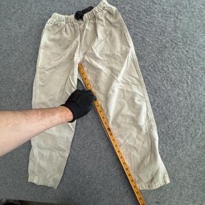 K&ouml;nnte beinhalten: Beigefarbene Hose mit elastischem Bund und integriertem schwarzen Schnalleng&uuml;rtel. Ein gelbes Ma&szlig;band liegt entlang der inneren Beinnaht und zeigt eine L&auml;nge von etwa 61 cm vom Schritt bis zum Saum an.